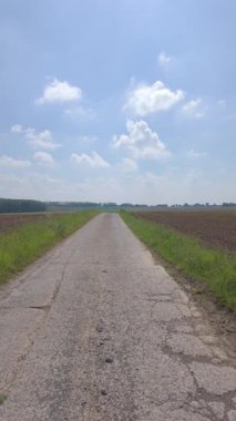 Kırık bir asfalt yol, beyaz bulutlu mavi gökyüzünün altında yeşil tarlalar boyunca esiyor, maceraya davetiye çıkarıyor.