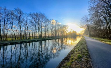 Sakin bir günbatımı, bereketli ağaçların çerçevelediği sakin kanal sularını ve gezmek için huzurlu bir patikayı yansıtır.