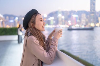 20 'li yaşlarda mini etekli genç bir Çinli kadın Hong Kong Adası' ndaki deniz kenarında bir grup gökdelene bakıyor.