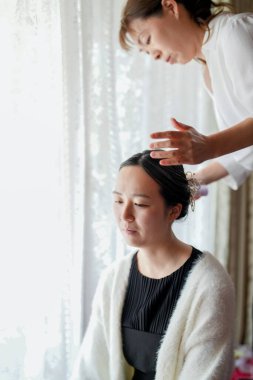 Bir kadın saçını başka bir kadına yaptırıyor. Saçını yaptıran kadın siyah bir elbise ve beyaz bir kazak giyiyor.