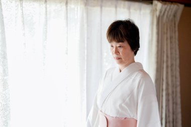 Beyaz kimonolu bir kadın pencerenin önünde duruyor. Pencereden dışarı bakıyor, muhtemelen dış dünyayı izliyor. Sakinlik ve sükunet kavramı