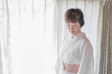 Beyaz kimonolu bir kadın pencerenin önünde duruyor. O, pembe bir kemer takıyor ve ellerini onun önünde sıkıca tutuyor. Oda loş bir şekilde aydınlatılmış ve perdeler çekilmiş.