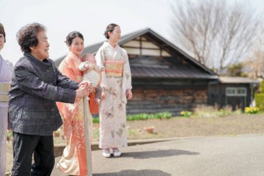Dışarıda bir grup insan bekliyor, içlerinden biri bebeği tutuyor. Sahnenin sıcak ve misafirperver bir atmosferi var. Yaşlı kadın bebeği kucağında tutuyor ve diğer insanlar etrafında dikiliyor.