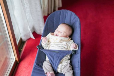 Bir bebek mavi bir yüksek sandalyede uyuyor. Sandalye kırmızı halıda. Bebek gülümsüyor ve üzerinde sarı bir gömlek var.