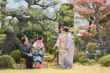 Geleneksel Japon giysileri içinde bir grup kadın bir bahçede oturuyor. Kadınlar gülümsüyor ve birbirlerinin arkadaşlığından zevk alıyor gibi görünüyorlar. Bahçe ağaçlar ve çalılarla dolu.