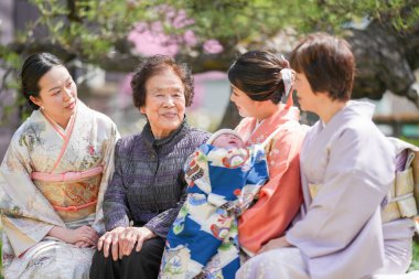 A group of women are sitting together, one of them holding a baby. Scene is warm and friendly, as the women are enjoying each other's company and sharing a moment together