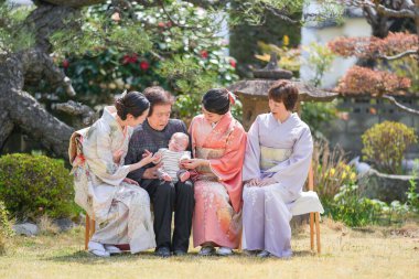 A group of people are sitting in a garden, with one of them holding a baby. The scene is peaceful and serene, with the people enjoying each other's company and the natural surroundings