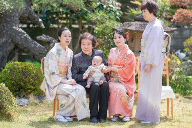 A group of people, including an elderly woman and a baby, are sitting in a garden. The woman is holding the baby, and the other people are sitting around them. The scene is peaceful and warm