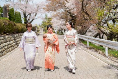 Three women in traditional Japanese clothing walk down a path. One of the women is carrying a baby