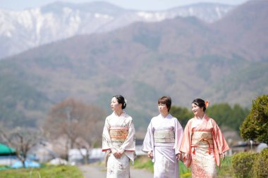Three women in traditional Japanese clothing are walking down a path in front of a mountain. The women are smiling and seem to be enjoying their time together