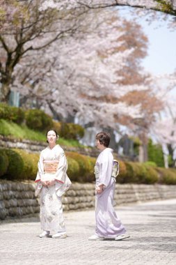 Two women in traditional Japanese clothing are walking down a path in a park. The women are smiling and seem to be enjoying their time together. The park is filled with cherry blossoms
