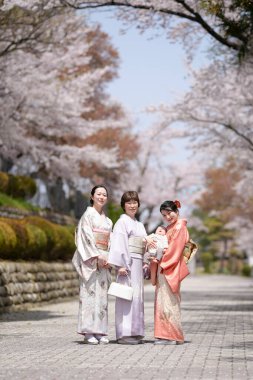 Three women in traditional Japanese clothing pose for a photo in front of cherry blossoms. Scene is serene and peaceful, as the women are surrounded by the beauty of nature