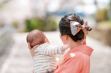 A woman is holding a baby in her arms. The baby is wearing a striped shirt. The woman is wearing a pink kimono