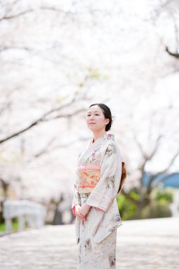 A woman in a kimono stands in front of a tree with pink blossoms. She is posing for a photo