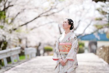 A woman in a kimono stands on a sidewalk in front of cherry blossoms. She is looking off into the distance, possibly lost in thought. Concept of tranquility and beauty