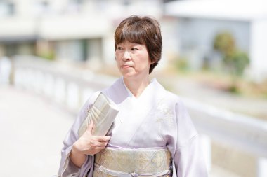 A woman in a kimono is holding a book. The scene is set on a bridge, with a white house in the background. The woman is in a contemplative mood, possibly reading the book or thinking about something