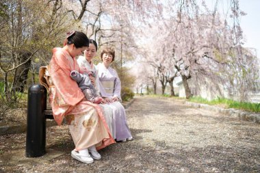 Three women in traditional Japanese clothing sit on a bench in a park. The women are smiling and seem to be enjoying each other's company. The park is filled with cherry blossoms