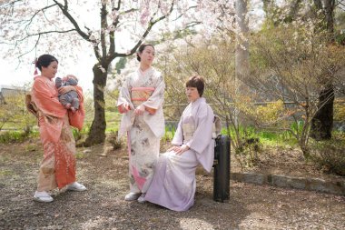Three women in traditional Japanese clothing are sitting on a bench in a park. One of them is holding a teddy bear. The scene is peaceful and serene, with the women enjoying each other's company