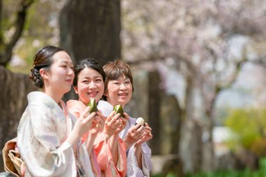 Three women are sitting on a bench, eating food and smiling. Scene is happy and relaxed