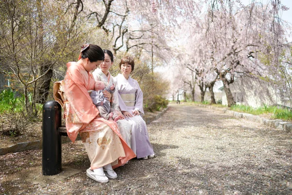 Three women in traditional Japanese clothing sit on a bench in a park. The women are looking at something, but it is not clear what they are looking at. The scene is peaceful and serene