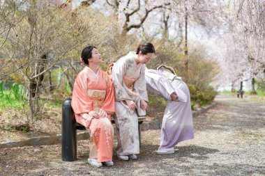 Geleneksel Japon elbiseli üç kadın bir parkta bir bankta oturuyor. Kadınlardan biri yerden bir şey almak için eğiliyor. Huzurlu ve huzurlu bir sahne.