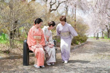 Geleneksel Japon elbiseli üç kadın bir yolda yürüyor. Kadınlardan biri bankta oturuyor.