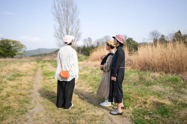 Üç kadın bir tarlada duruyor, biri siyah elbise giyiyor. Uzakta bir şeye bakıyorlar.