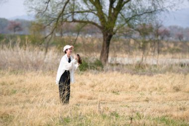 Beyaz elbiseli bir kadın elinde bir kuşla bir tarlada duruyor. Sahne huzurlu ve huzurlu. Kadın ve kuş görüntüde yaşayan tek canlılar.