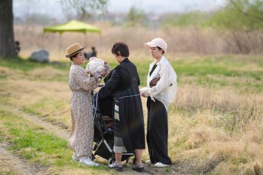 Üç kadın bir tarlada duruyor, biri bebek tutuyor. Diğer iki kadın onun arkasında duruyor ve hepsi şapka takıyor. Sahne rahat ve rahat.