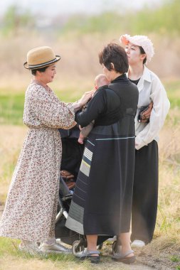 Üç kadın bir tarlada duruyor, biri bebek tutuyor. Diğer iki kadın onun arkasında duruyor ve hepsi şapka takıyor. Olay yeri sıradan ve dostane.
