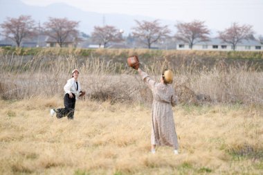 Bir tarlada beyzbol oynayan iki kadın. Biri koşuyor, diğeri de top atıyor. Sahne neşeli ve neşeli.