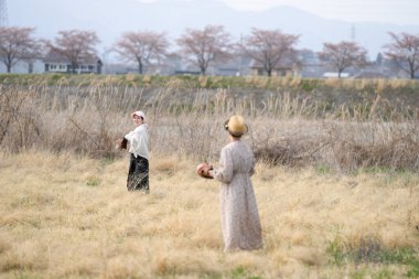 İki kadın bir tarlada duruyor, biri beyzbol sopası tutuyor. Tarla kuru ve çorak, görünürde ağaç yok. Manzara ıssız ve ıssız.