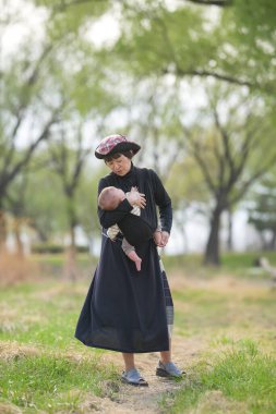 Bir kadın kucağında bir bebek tutuyor. Bebek siyah bir kıyafet giyiyor. Kadın siyah bir elbise ve bir şapka giyiyor. Sahne arka planda ağaçlar olan bir parkta geçiyor.
