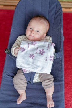 Üzerinde beyaz bir battaniye olan mavi bir sandalyede oturan bir bebek var. Bebek gülümsüyor ve kameraya bakıyor.