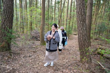 Kollarında bir bebekle ormanda yürüyen bir kadın. Bebek şapka takıyor.