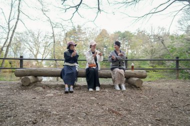 Parkta kütüğün üzerinde oturan üç kadın. İçlerinden biri bir şişe tutuyor. Manzara huzurlu ve rahatlatıcı.