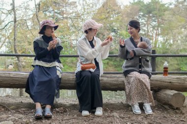 Üç kadın kütüğün üzerinde oturuyor, biri bebek tutuyor. Manzara huzurlu ve rahatlatıcı.