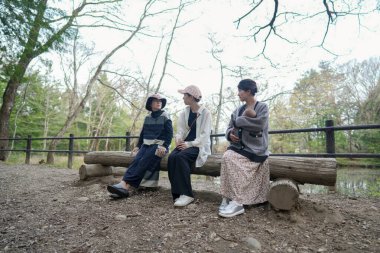 Parktaki bir kütüğün üzerinde üç kişi oturuyor. İçlerinden biri bir bebek tutuyor. Manzara huzurlu ve rahatlatıcı.