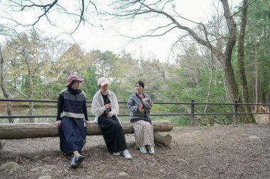 Parkta kütüğün üzerinde oturan üç kadın. İçlerinden biri pembe bir şapka takıyor. Bir şişeden içiyorlar.