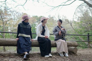Üç kadın parkta bir kütüğe oturmuş, içlerinden biri bebek tutuyor. Manzara huzurlu ve rahatlatıcı, çünkü kadınlar doğal ortamda birbirlerine eşlik etmekten hoşlanıyorlar.