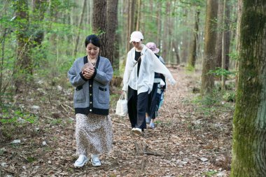 Bir kadın iki kişiyle birlikte ormanda yürüyor. Elinde bir cep telefonu tutuyor.