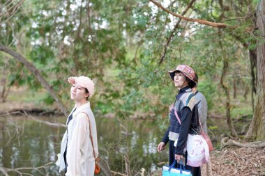 İki kadın bir nehrin kenarında duruyor, biri pembe şapkalı. El çantaları taşıyorlar ve açık havanın tadını çıkarıyorlar.