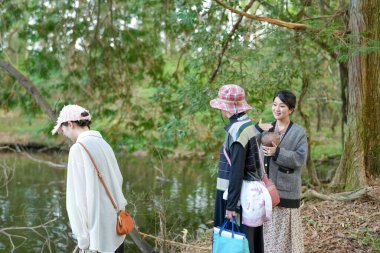 Bir grup insan, elinde köpek tutan bir kadınla nehrin kenarında duruyor. Sahne huzurlu ve sakin, arka planda suyun sesi ve ağaçlar var.