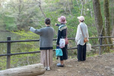 Üç kadın göletin kenarında bir patikada duruyorlar, uzaktaki bir şeye bakıyorlar. Şapka takıyorlar ve çanta taşıyorlar.