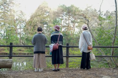 Üç kadın, göle bakan bir köprüde duruyor. Hepsi şapka takıyor ve çanta taşıyor. Kadınlar göletin doğal güzelliğinin tadını çıkarırken manzara huzurlu ve huzurlu.