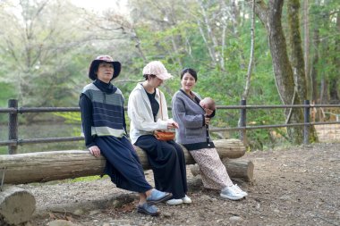 Üç kadın parkta bir kütüğe oturmuş, içlerinden biri bebek tutuyor. Manzara huzurlu ve rahatlatıcı, çünkü kadınlar birbirlerinin arkadaşlığından ve doğal çevreden hoşlanıyorlar.