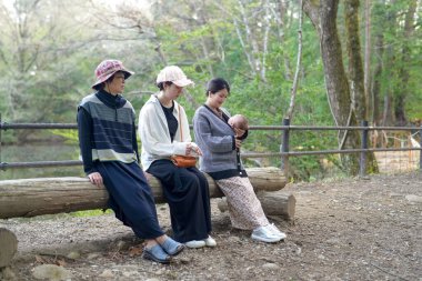 Bir grup kadın parkta bir kütüğe oturmuş, içlerinden biri bebek tutuyor. Manzara huzurlu ve rahatlatıcı, çünkü kadınlar birbirlerinin arkadaşlığından ve doğal çevreden hoşlanıyorlar.