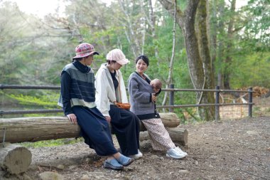 Bir grup kadın ormandaki bir kütüğe oturmuş, içlerinden biri bebek tutuyor. Manzara huzurlu ve huzurlu, çünkü kadınlar birbirlerine eşlik etmekten ve doğal çevreden hoşlanıyorlar.