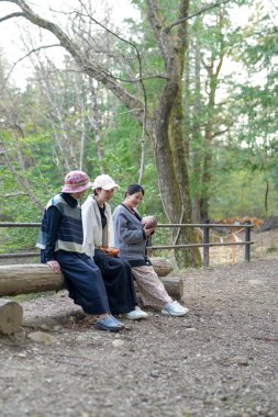 Üç kadın ormandaki bir kütüğün üzerinde oturuyor. İçlerinden biri bir bebek tutuyor.