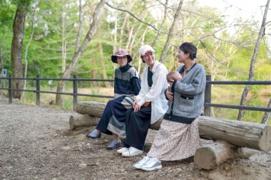 Üç kadın parkta bir kütüğe oturmuş, içlerinden biri bebek tutuyor. Manzara huzurlu ve rahatlatıcı, çünkü kadınlar doğal ortamda birbirlerine eşlik etmekten hoşlanıyorlar.